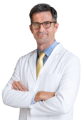VTC_DrKindelan_Cutout_optimized Dr. Joshua Kindelan is captured in a professional portrait wearing a white lab coat over a blue plaid dress shirt and a yellow tie. He is standing with his arms crossed in a clinical environment.